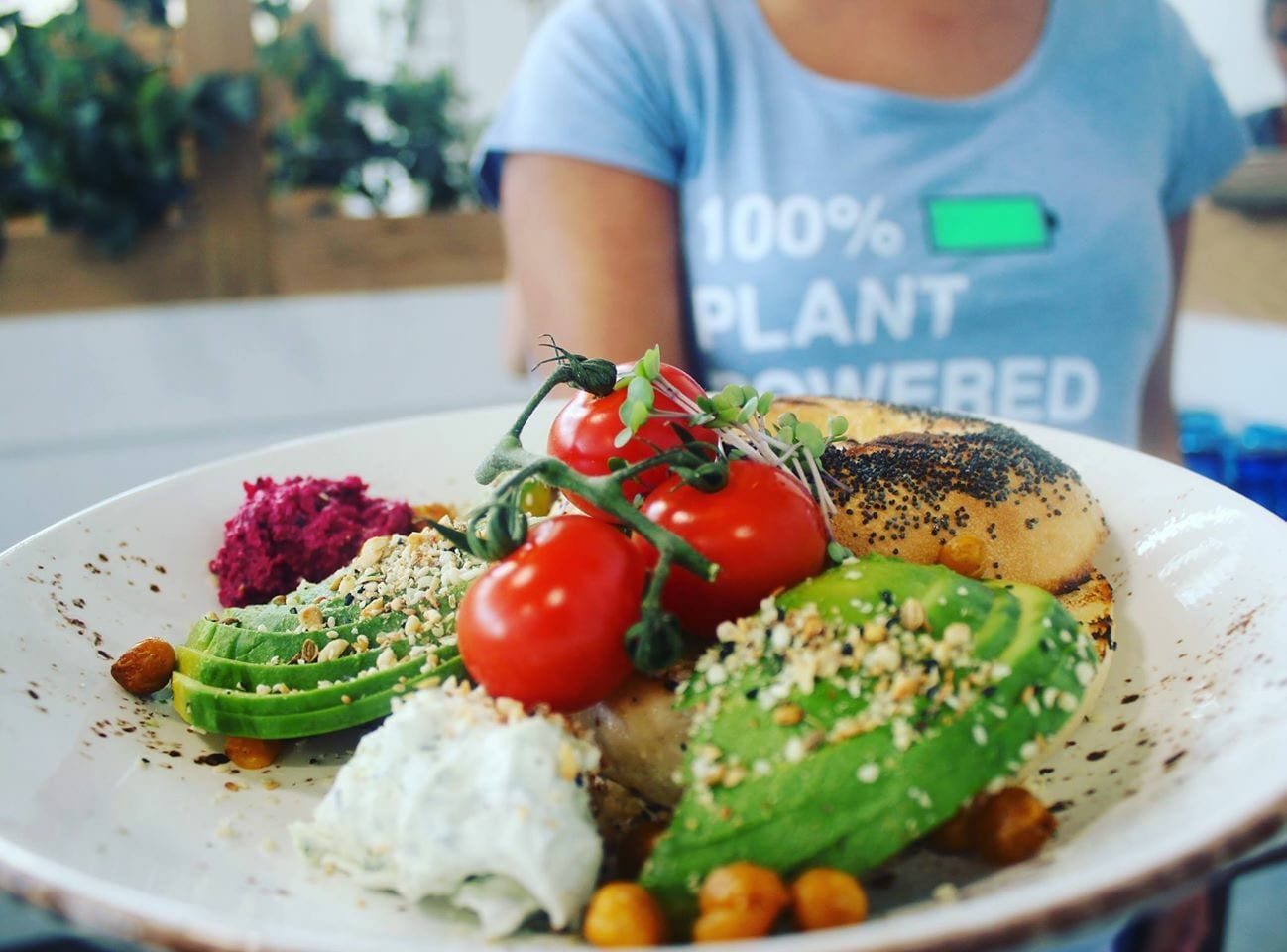 Plant-based breakfast plate with avocado, cherry tomatoes, beetroot hummus and bagel, served by staff in a 100% Plant Powered t-shirt