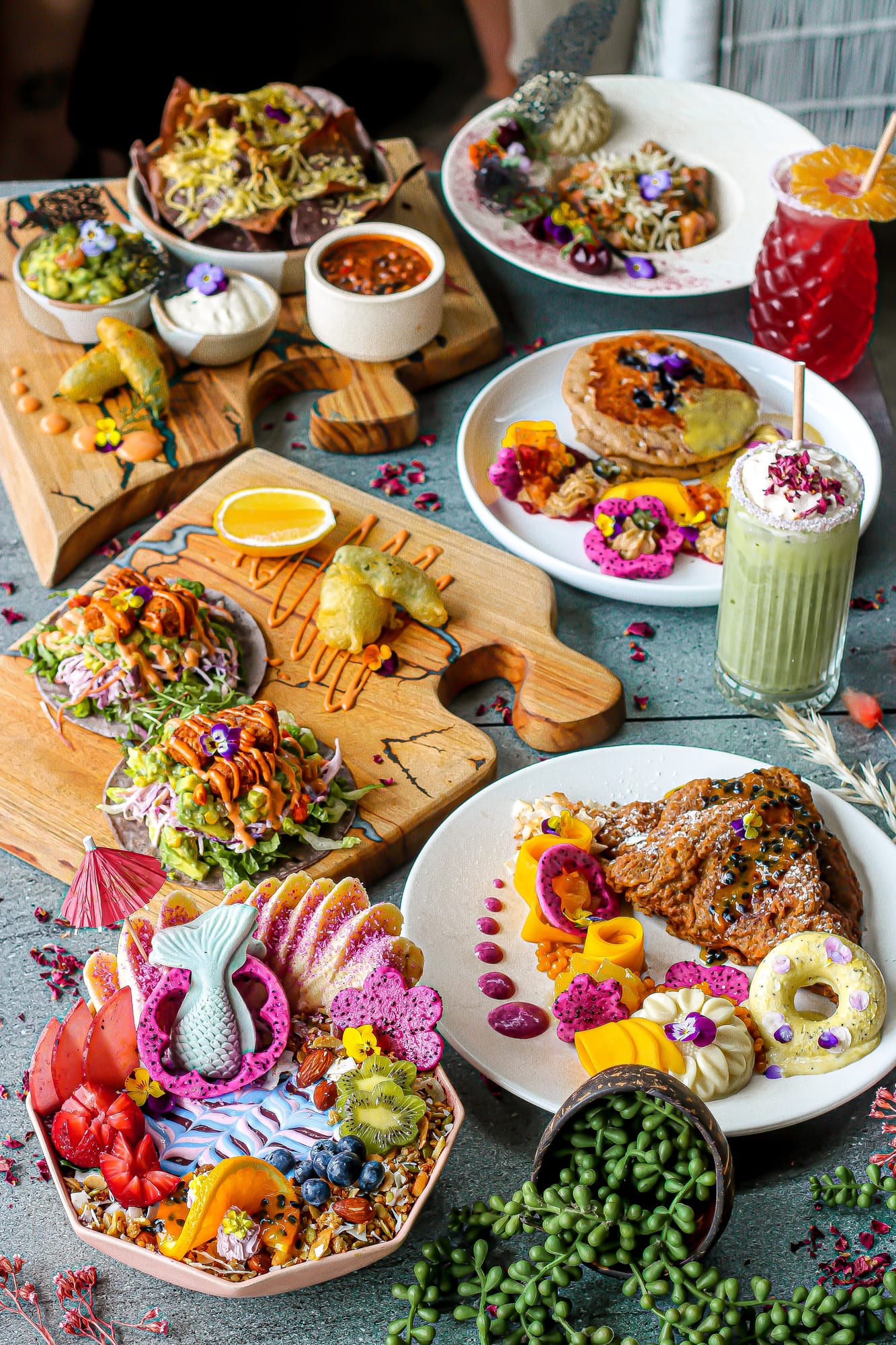 Overhead spread of colourful plant-based dishes, smoothie bowls, tacos, pancakes and tropical drinks at The Cardamom Pod